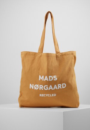 Braune Stofftasche mit zwei Griffen, Vorderseite mit weißem Schriftzug "Mads Nørgaard Recycled", auf einem weißen Podest platziert.