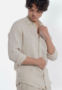 Beige linnen shirt met lange mouwen, opgerold bij de ellebogen, knoopsluiting aan de voorkant, zachte textuur en subtiele stiknaaddetails.
