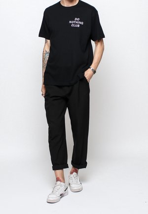 Person trägt schwarzes "DO NOTHING CLUB" T-Shirt, schwarze hochgekrempelte Hose, weiße Sneakers, zeigt tätowierten Unterarm und Knöchel.