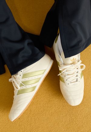 Personne portant des baskets Adidas blanches et dorées avec un pantalon noir, debout sur un tapis jaune moutarde.