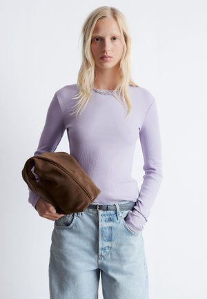 Helles lila, geripptes Langarmoberteil, getragen zu hellblauen Jeans. Hält eine Tasche aus braunem Wildleder. Einfaches Design, keine Muster oder auffälligen Beschläge.