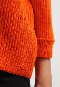 Gestrickter orangefarbener Pullover mit gerippter Textur, der einen umgeschlagenen Saum und ein kleines, oranges Logo-Detail nahe dem Saum aufweist.