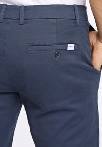 Navy blue chinos made of cotton with a smooth texture, featuring a rear pocket with a button closure and a small brand label.