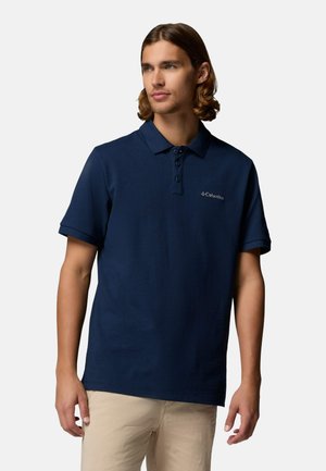 Junger Mann mit schulterlangem Haar, der ein navyblaues Columbia-Poloshirt und beige Hosen trägt und leicht zur Seite schaut.