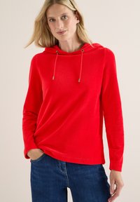 Femme blonde portant un sweat à capuche rouge vif à manches longues et un jean bleu foncé, se tenant avec une main dans la poche.