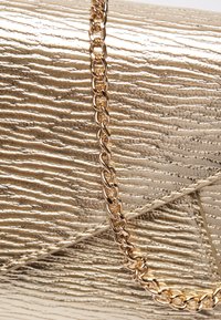 Metalická zlatá kabelka typu clutch s texturovaným, vlnitým povrchem. Obsahuje zlatý řetízkový popruh a trojúhelníkový klopový design, který dodává moderní vzhled.