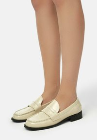 Cream patent leather loafers with a square toe and low black heel, featuring a smooth finish and subtle stitching details.