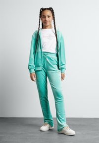 Menta zelena velur trenirka z zip up kapuco in hlačami na vrvico, kombinirana z belo majico z logojem iz umetnih kamnov. Bele superge.