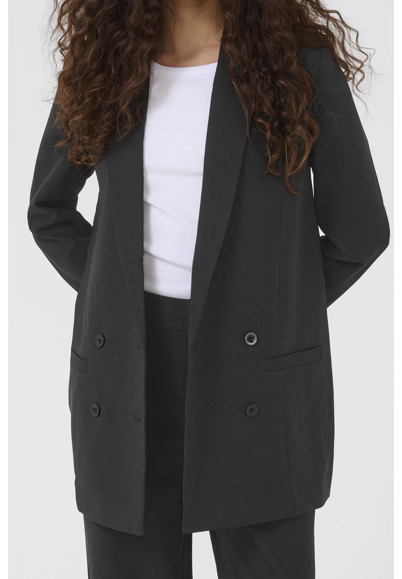 Femme portant un long blazer noir croisé et un pantalon noir, avec un chemisier blanc côtelé en dessous, les mains derrière le dos.