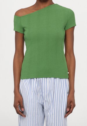 Frau trägt ein grünes kurzärmeliges schulterfreies Oberteil und blau-weiß vertikal gestreifte Hose, steht vor einem einfachen Hintergrund.