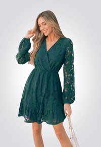 Abito verde in pizzo con maniche lunghe, scollatura a V e orlo svasato. Presenta motivi floreali e una vita aderente, abbinato a una borsa piccola.
