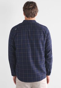 Chemise à manches longues à carreaux bleu marine en coton doux, avec un col boutonné et un logo visible sur la poitrine. Présente un ourlet arrondi et des poignets boutonnés.