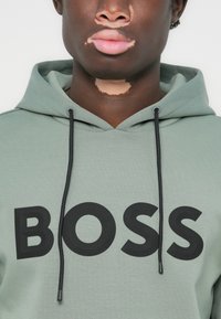 Sudadera de color verde claro con el logo negro "BOSS", que cuenta con una capucha con cordón, textura suave y un ajuste relajado.