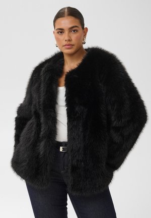 Femme aux cheveux foncés portant une grande veste en fausse fourrure noire sur un haut blanc et un jean foncé, debout devant un fond gris uni.