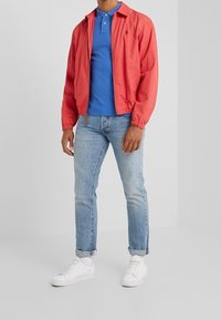 Röd lättviktig jacka, blå polo skjorta, ljusblå jeans och vita sneakers. Jackan har elastiska ärmslut och dragkedjestängning. Avslappnad passform.