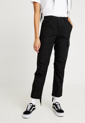 Woman wearing black cargo pants with front flap pockets, white crew socks, black and white sneakers, and a white short-sleeve shirt, standing.