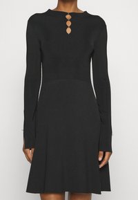 Robe en maille noire à manches longues, texture côtelée, corsage ajusté, jupe évasée, avec trois découpes ovales au niveau de l'encolure arrière.