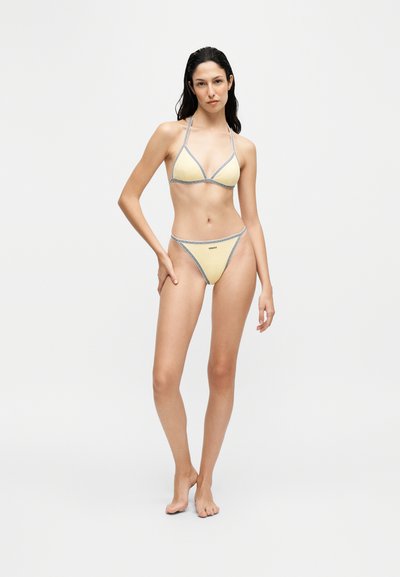 Femme aux cheveux foncés portant un bikini jaune avec une bordure grise, debout pieds nus devant un fond clair uni.