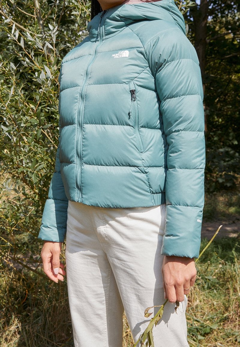 Personne portant une doudoune North Face bleu sarcelle et un pantalon blanc, tenant une petite plante séchée, debout en plein air près d'un feuillage vert.