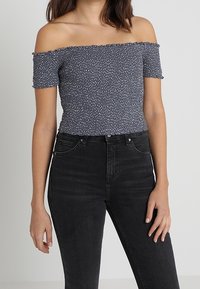 Top azul marino, sin mangas y con un diseño fruncido, de mangas cortas. Combinado con jeans negros de tiro alto. La textura está ligeramente recogida.