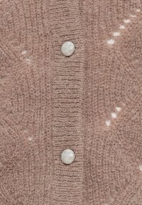 Heled pink villakardigan pehmea tekstuuriga viimistlusega, millel on kolm ümmargust, kalliskividega nööpi ja keeruline muster.