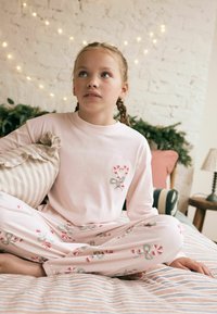 Vaaleanpunainen yöpuku, jossa on sydän- ja karkkitangon kuvio, sisältää pyöreäkaulaisen paidan ja elastiset housut. Raidallinen tyyny lepää lähellä.