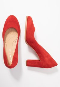 Escarpins à talons hauts en daim rouge avec un bout arrondi et un talon épais. La semelle intérieure est perforée pour un confort accru.