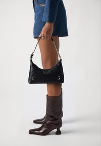 Borsa nera in ecopelle con motivo in rilievo, hardware in tonalità argento e tracolla a catena, tenuta in una mano accanto a stivali alla caviglia in pelle marrone.