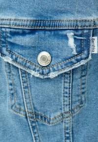 Ljusblå denimjacka med en sliten frampå ficka, silverfärgad tryckknapp och kontrasterande orange sömnadsdetaljer. Märket är märkt "Bershka."
