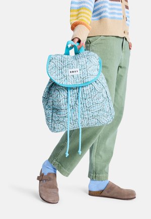 Persoon in groene broek en een multikleurige gestreepte trui die een blauw Roxy-rugzak met bloemmotief en trekkoordsluiting vasthoudt.