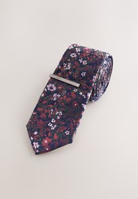 Námořnicky modrá kravata s květinovým vzorem v odstínech růžové a zelené. Hladká látka s špičatým koncem a připevněnou stříbrnou sponou na kravatu.