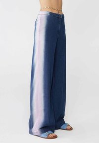 Pantaloni in denim a gamba larga con una sfumatura che va dal blu sbiadito al rosa, con una texture liscia e chiusura frontale con bottone. Abbinati a sandali blu.