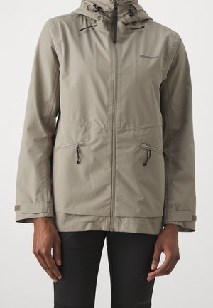 Světlé béžové kapucové sako se zapínáním na přední zip, dvěma zipovanými bočními kapsami, nastavitelnými pásky na zápěstí, nošené stojící osobou.