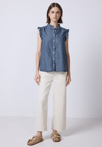 Top senza maniche in denim scuro con scollatura arricciata e chiusura a pulsante, abbinato a pantaloni beige chiaro a gamba larga e sandali infradito color nocciola.