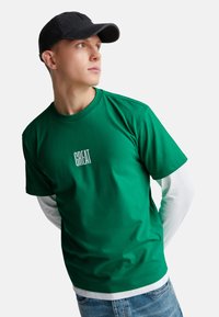 Camiseta de algodón verde superpuesta sobre una camisa de manga larga blanca, con un logo "GREAT" centrado. Combinada con jeans azules y una gorra negra.