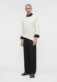 Homme portant un pull en maille torsadée blanc sur une chemise noire à col, un pantalon noir à jambes larges et des sandales noires, debout devant un fond uni.