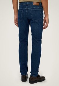 Vaqueros de mezclilla de lavado oscuro con un ajuste slim, que cuentan con dos bolsillos traseros y una etiqueta de cuero en la cintura, combinados con zapatos oscuros.