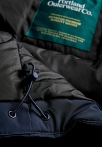 Czarna izolowana kurtka z teksturowanej tkaniny, z zapięciem na sznurki i zieloną etykietą marki wszytą od wewnątrz.