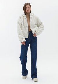 Felpa bianca e soffice con zip e cappuccio, abbinata a un dolcevita marrone chiaro, jeans scuri a vita alta e sneakers chiare.