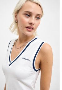 White sleeveless top with a v-neck, navy trim, and "iets frans..." embroidery on the left side. Cotton fabric, fitted silhouette.