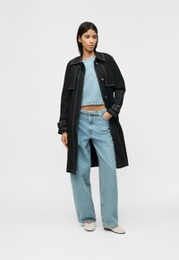 Γυναίκα που στέκεται φορώντας μαύρο καμπαρντίνα πάνω από ανοιχτό μπλε κοντό πουλόβερ και τζιν με φαρδιά πόδια, με λευκά παπούτσια slip-on.