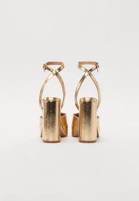 Sandali alti metallici dorati presentano un tacco spesso, una cinghia alla caviglia con fibbia e un design della punta scolpito, mostrando una texture liscia.