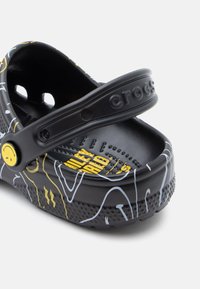 Crocs noirs à finition brillante, ornés de motifs linéaires blancs et jaunes, avec une semelle moulée et une sangle proéminente portant le nom de la marque.