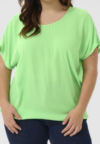 Personne grande taille portant un haut à manches courtes ample de couleur vert vif et un jean bleu foncé, les mains reposant près des hanches.