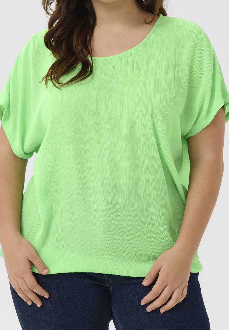 Personne grande taille portant un haut à manches courtes ample de couleur vert vif et un jean bleu foncé, les mains reposant près des hanches.