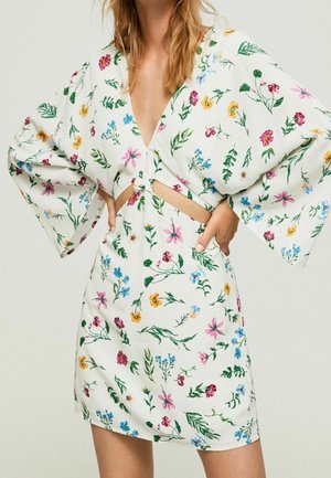Vrouw draagt een witte jurk met kleurrijke bloemenprints, wijde mouwen, een diepe V-hals en een uitgesneden gedeelte aan de voorkant bij de taille.