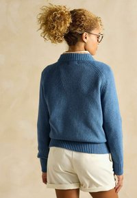 Pulover tricotat albastru cu guler și tiv cu nervuri, având mâneci raglan și o textură unică, asortat cu șorturi de culoare deschisă.