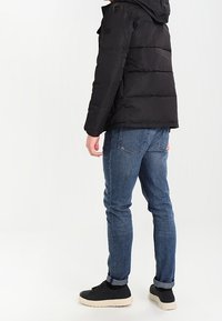 Svart vadderad jacka med huva, kombinerad med blå denimjeans och svarta sneakers med en krämfärgad sula. Enkelt och funktionellt design.