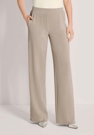 Femme portant un pantalon large beige taille haute avec poches latérales, associé à des chaussures blanches à bout pointu sur un fond uni.
