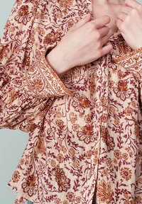 Blouse à motifs floraux en pêche clair avec des accents bordeaux et dorés. Présente une fermeture à boutons, de longues manches bouffantes et des poignets détaillés.